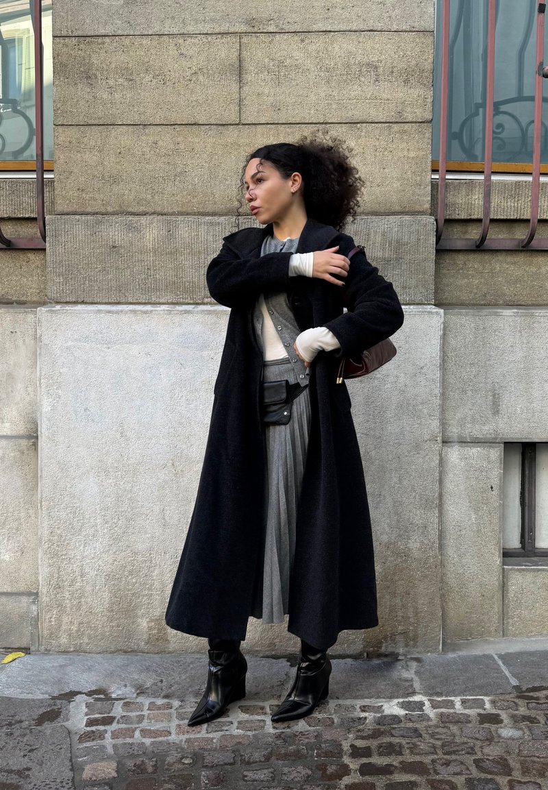 Kvinde med krøllet hår iført en lang sort frakke, grå plisseret nederdel, sorte støvler og holder en brun skuldertaske, stående på brostensbelægning.