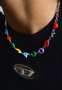 Encolure d'un haut noir côtelé avec une découpe ovale et un fermoir argenté, ornée d'un collier de perles en céramique multicolores sur chaîne.