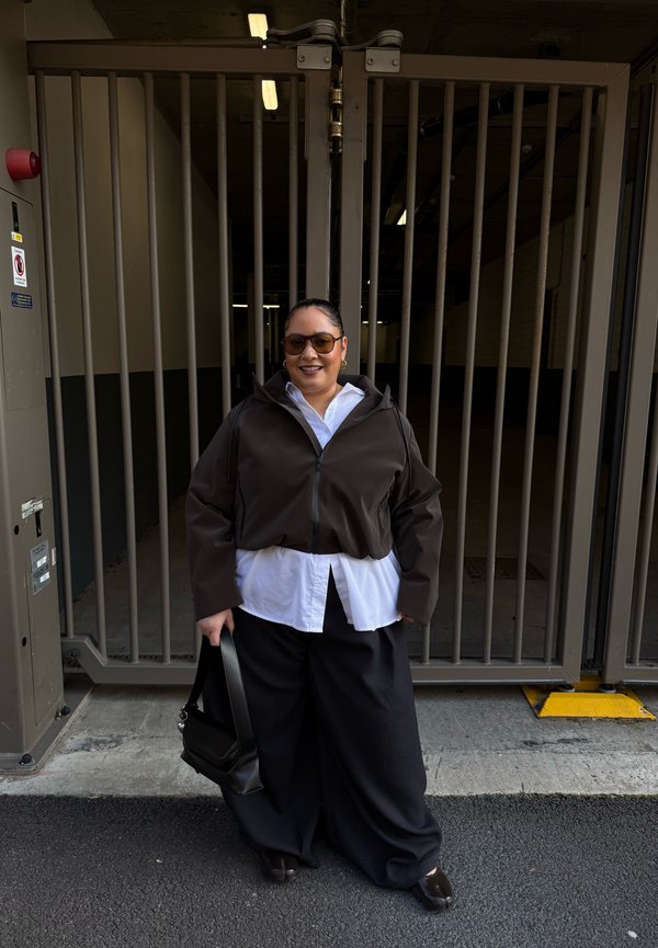 Femme portant des lunettes de soleil, veste marron sur chemise blanche, pantalon noir large, tenant un sac à main noir, debout devant une grille de sécurité en métal.