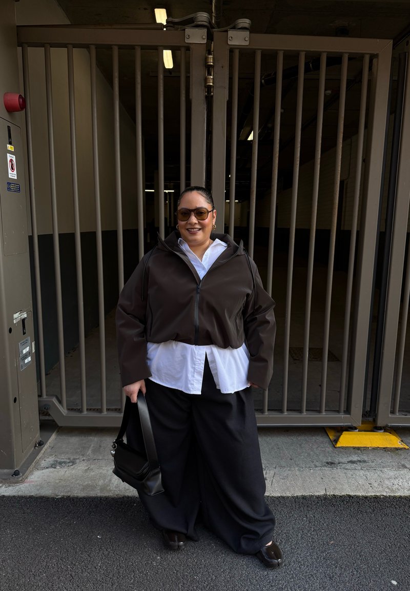 Femme portant des lunettes de soleil, veste marron sur chemise blanche, larges pantalons noirs, tenant un sac à main noir, debout devant une grille de sécurité en métal.