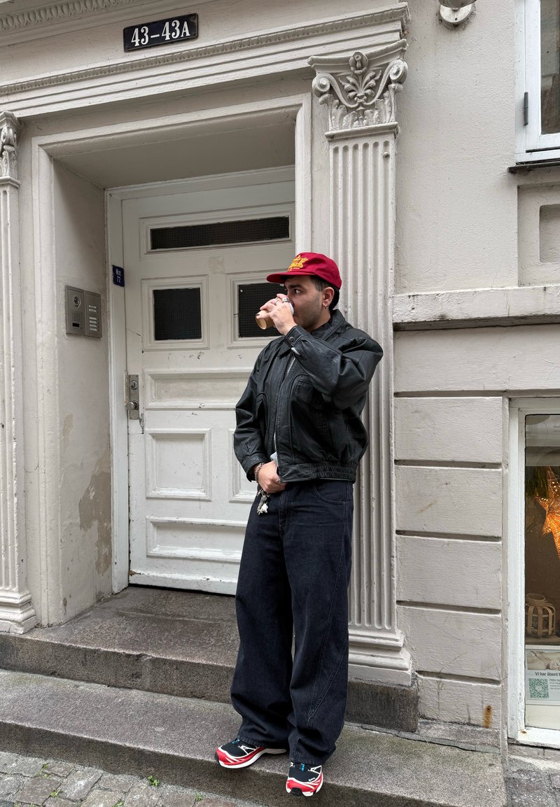 Un homme portant une veste en cuir noire, un jean ample sombre et une casquette rouge se tient près d'une porte blanche, tenant un verre avec un porte-clés visible.