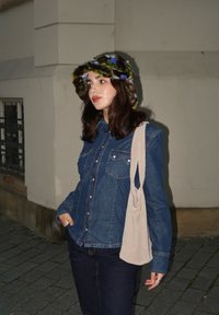 Τζιν πουκάμισο με λεπτομέρειες από κουμπιά, σκούρα τζιν και ένα πολύχρωμο καπέλο από γούνα. Μία ελαφριά τσάντα ώμου κρέμεται από τον έναν ώμο. Νυχτερινό σκηνικό.