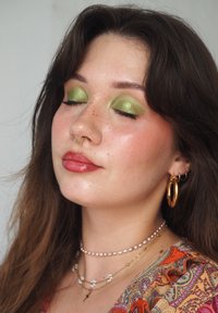 Nahaufnahme einer Person mit gold-grünem Lidschatten, einem goldenen Septumring und mehreren Halsketten. Die Haut hat Sommersprossen und einen sanften Schimmer.