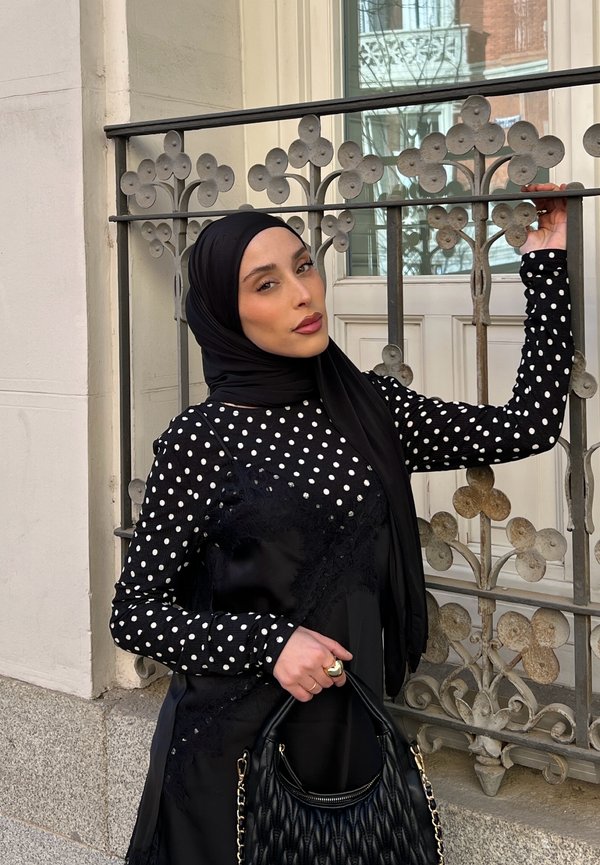 Femme en hijab noir et haut à pois pose avec un sac à main noir matelassé devant une rambarde en fer décorative en extérieur.