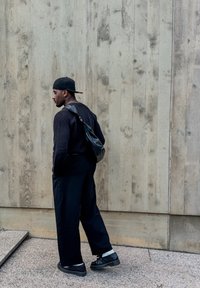 Maglione nero a maniche lunghe, pantaloni neri larghi, zaino nero con una tracolla, scarpe nere, in piedi contro una parete di legno chiaro e texturizzato.