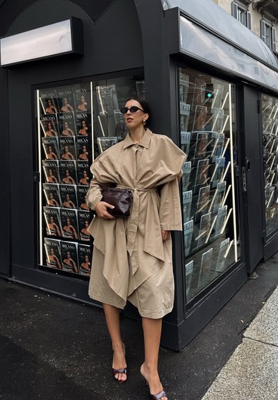 Gabardina beige de gran tamaño con cuello dramático y dobladillo asimétrico, combinada con un bolso de mano marrón y gafas de sol oscuras, frente a un expositor de revistas.