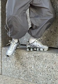 Las zapatillas Nike plateadas cuentan con un upper texturizado, un diseño curvado, una suela gruesa con amortiguación de aire visible y acentos negros. Combinadas con pantalones grises.