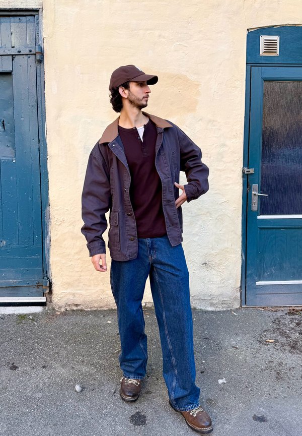 Un homme portant une casquette, une veste de travail, une chemise sombre et un jean ample pose entre deux portes bleues en regardant sur le côté.