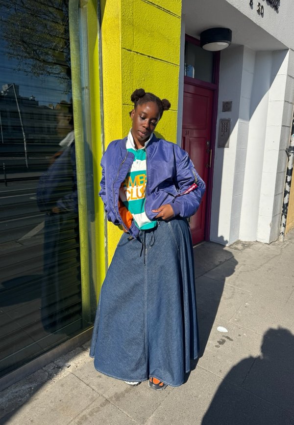 Joven con peinado de dos moños se apoya en una pared amarilla, lleva puesta una chaqueta azul, camisa a rayas, falda larga vaquera y zapatillas en una acera de la ciudad.