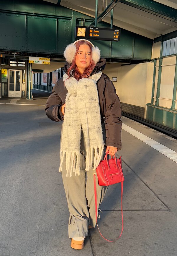 Zwarte pufferjack, grijze broek met trekkoord, witte gestructureerde sjaal met franjes, rode handtas, beige schoeisel, buitenomgeving van een treinstation.