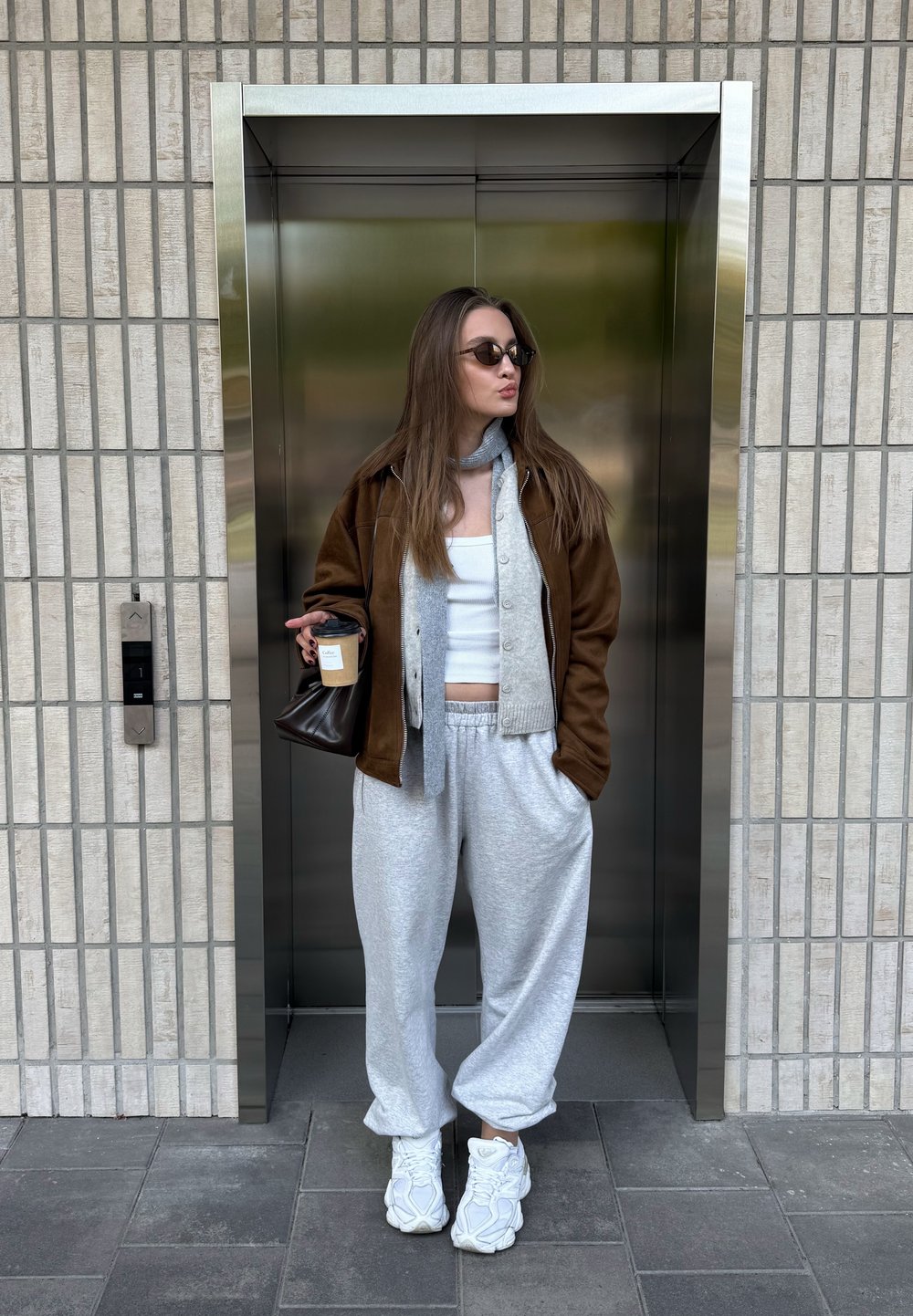 Verzameling van een bruine jas over een grijze cardigan en een witte tanktop, gecombineerd met grijze sweatpants en witte sneakers. Houdt een donkere tas en een koffiekuip vast.