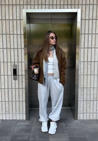 Verzameling van een bruine jas over een grijze cardigan en een witte tanktop, gecombineerd met grijze sweatpants en witte sneakers. Houdt een donkere tas en een koffiekuip vast.