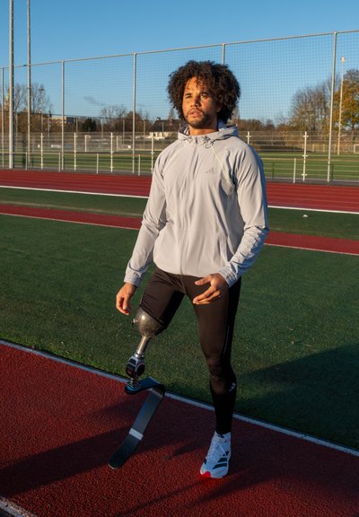 Atleta con chaqueta deportiva gris claro y mallas negras, con una prótesis de pierna de fibra de carbono y zapatillas blancas con detalles en rojo.