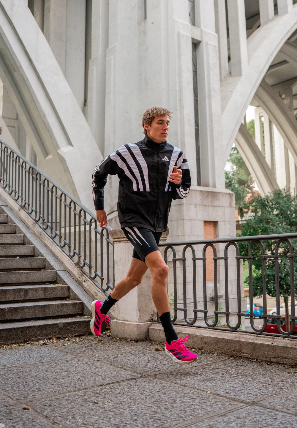 Jovem a correr a descer uma colina perto de arcos urbanas de concreto, vestido com roupa desportiva preta e sapatilhas de corrida cor-de-rosa brilhante.