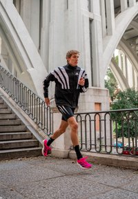 Jovem a correr a descer uma colina perto de arcos urbanas de concreto, vestido com roupa desportiva preta e sapatilhas de corrida cor-de-rosa brilhante.