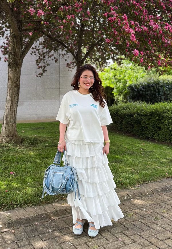 Lächelnde Frau mit langen Haaren trägt weißen mehrlagigen Rock, übergrosses weisses T-Shirt, blaue Ballerinas und hält eine Jeans-Handtasche unter einem Baum mit pinken Blüten im Freien.