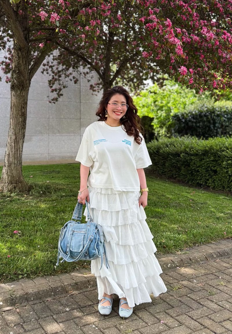 Femme souriante aux longs cheveux portant une jupe blanche à volants, un t-shirt blanc oversized, des ballerines bleues, et tenant un sac à main en jean sous un arbre à fleurs roses à l'extérieur.