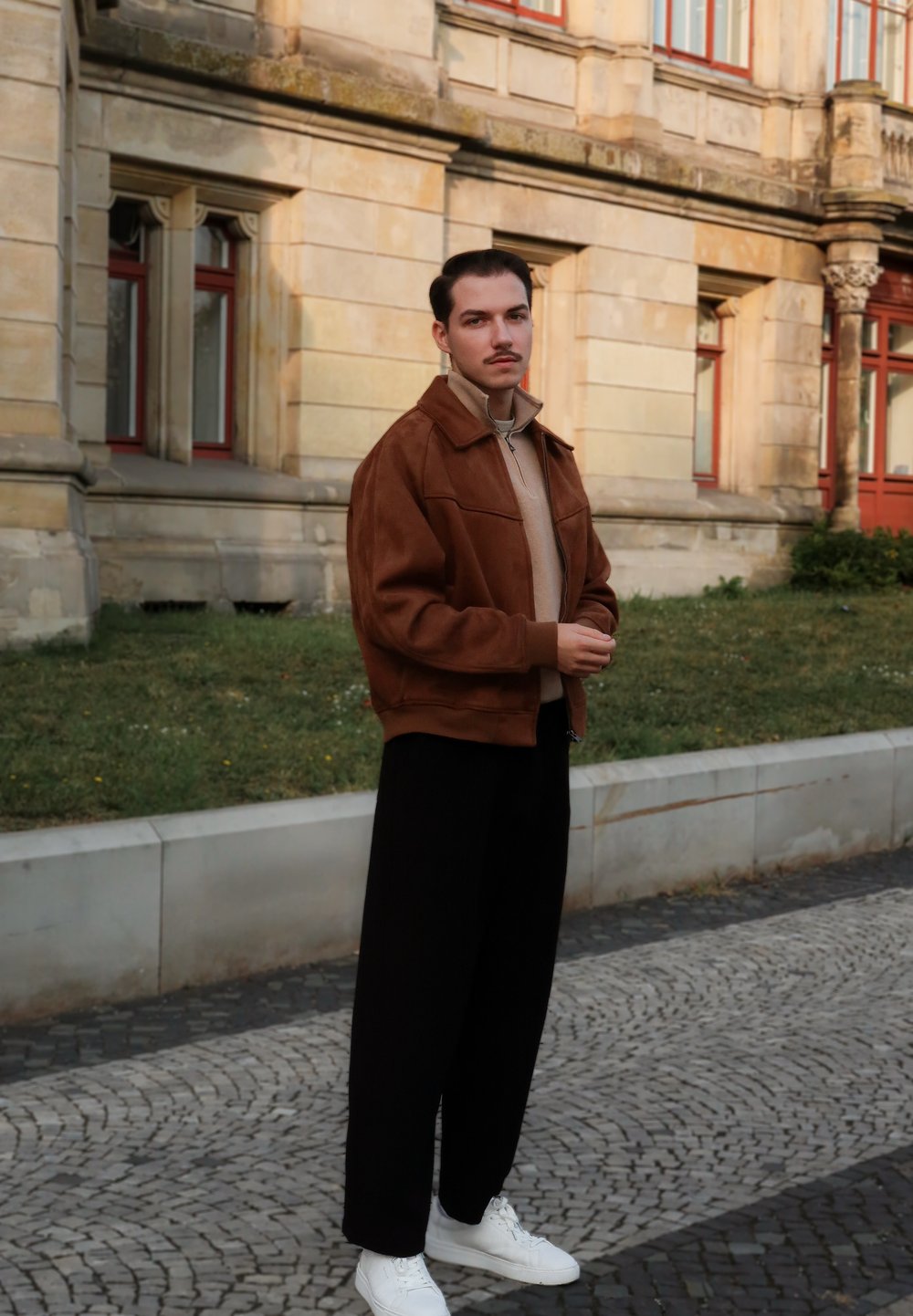 Brun ruskindsjakke, beige turtleneck, sorte bukser og hvide sneakers. Model står udendørs med grå stenbelægning og græs i baggrunden.