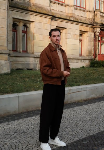 Chaqueta de ante marrón, suéter de cuello alto beige, pantalones negros y zapatillas blancas. El modelo está de pie al aire libre con un fondo de pavimento de piedra gris y hierba.