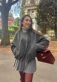 Γκρι υπερμεγέθης μπουφάν bomber με ψηλό γιακά, συνδυασμένο με γκρι πλισέ φούστα και μπορντό τσάντα. Το φόντο είναι γεμάτο από φθινοπωρινά φύλλα και δέντρα.