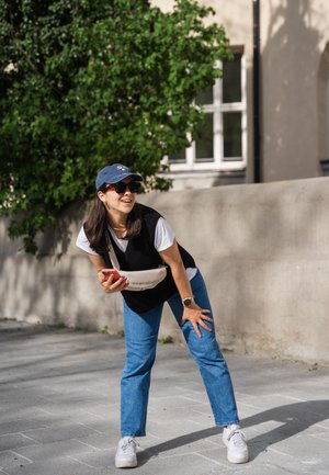 Suéter negro sin mangas sobre una camiseta blanca, jeans azules y zapatillas blancas. Un bolso cruzado beige y una gorra azul marino. Entorno exterior brillante.