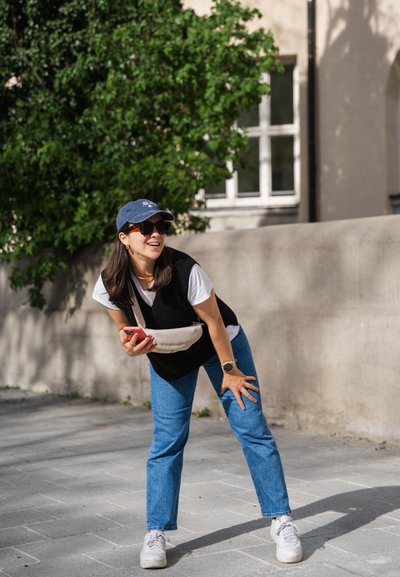 Suéter negro sin mangas sobre una camiseta blanca, jeans azules y zapatillas blancas. Un bolso cruzado beige y una gorra azul marino. Entorno exterior brillante.