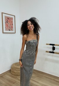 Grauer Denim-Maxi-Kleid mit tailliertem Oberteil, definierten Nähten und einem einzelnen Schulterträger, vor einem minimalistischen Hintergrund.