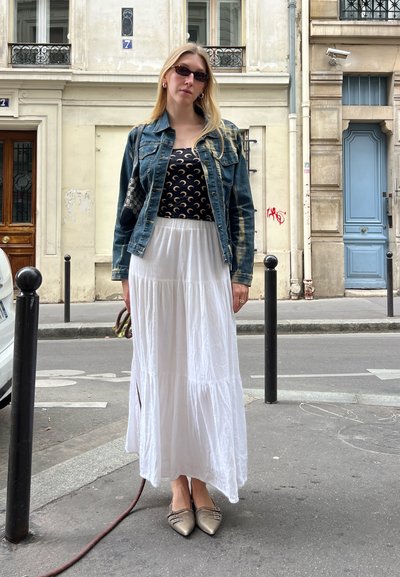 Chaqueta de mezclilla sobre una blusa negra con estampado, combinada con una falda maxi blanca. Incluye zapatos planos plateados y un bolso con estampado.