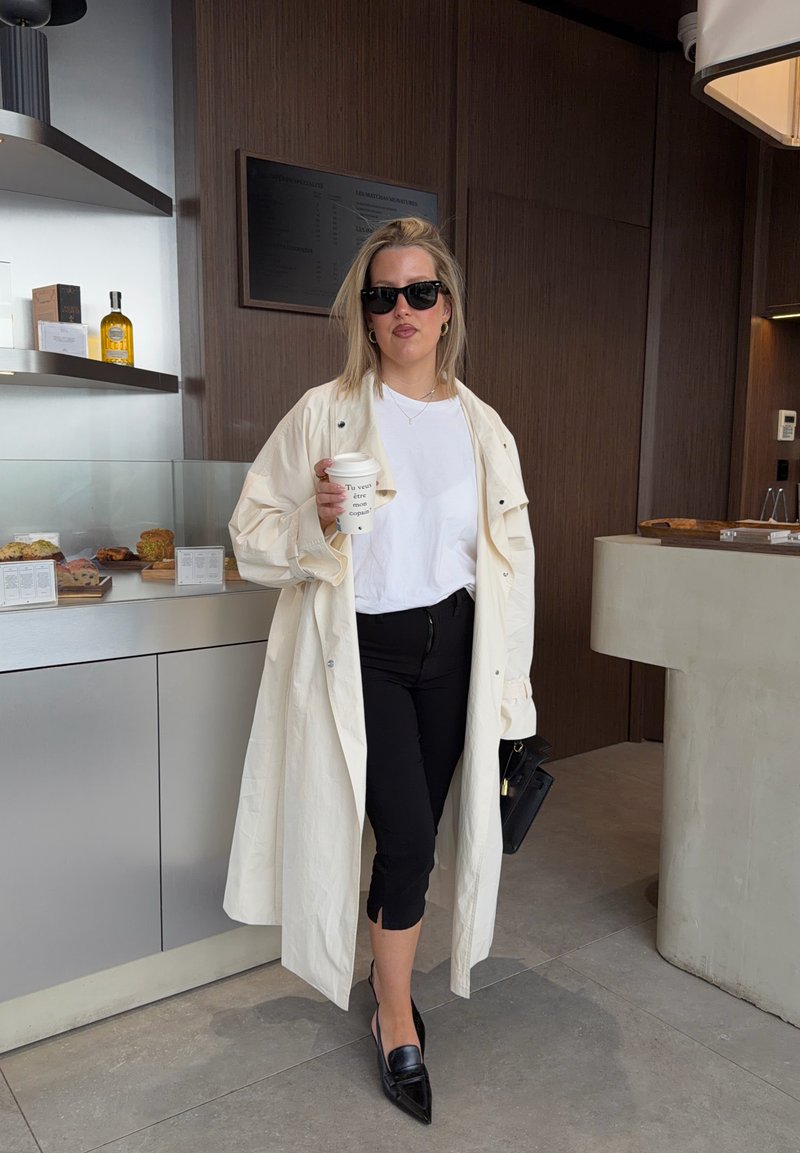 Femme portant des lunettes de soleil, un long manteau blanc, une chemise blanche et un pantalon noir, tenant une tasse de café dans un intérieur de café moderne.