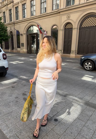 Top corto blanco y falda midi a juego, ambos hechos de una tela suave. Llevando un gran bolso verde oliva con un patrón floral. Sandalias negras.
