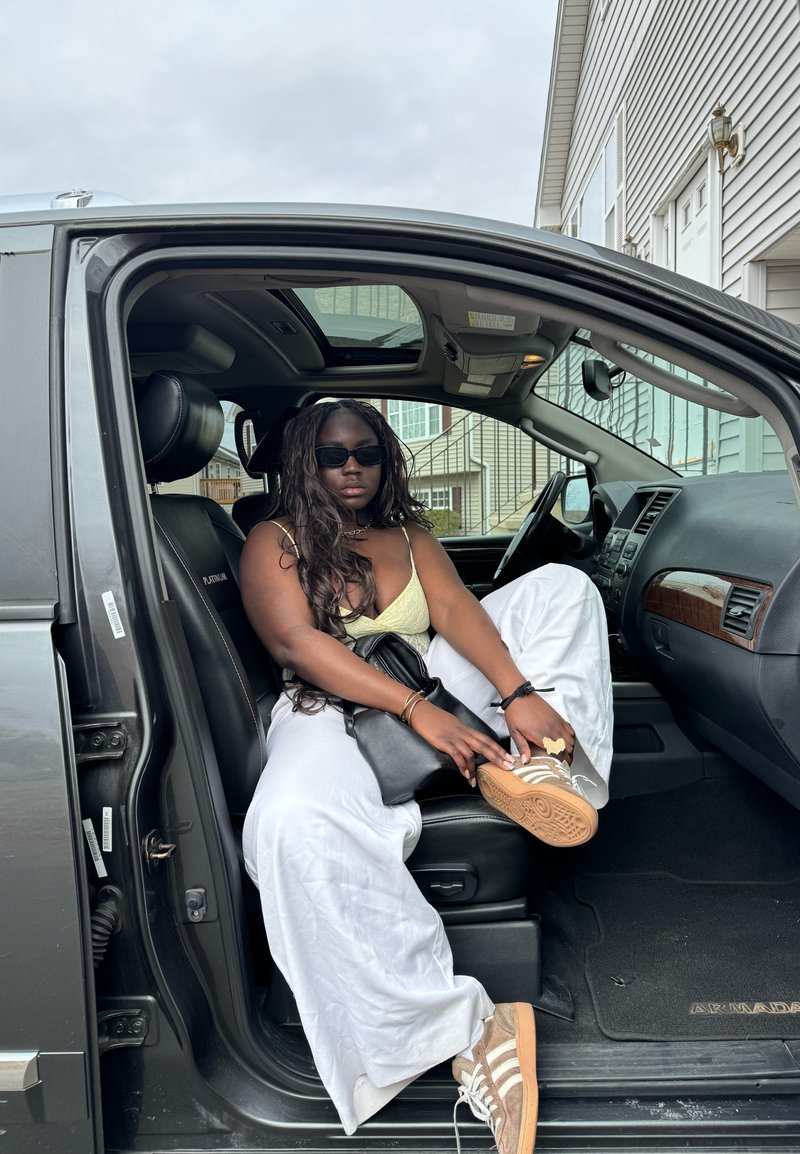 Femme portant des lunettes de soleil, un haut jaune et un pantalon blanc, assise sur le siège avant ouvert d’un SUV noir, ajustant sa chaussure avec des maisons visibles à l’extérieur.