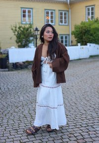 Bruine oversized jas, witte maxi-jurk met bloemaccenten en bruine sandalen. Stenen bestrating en een achtergrond van houten gebouwen.