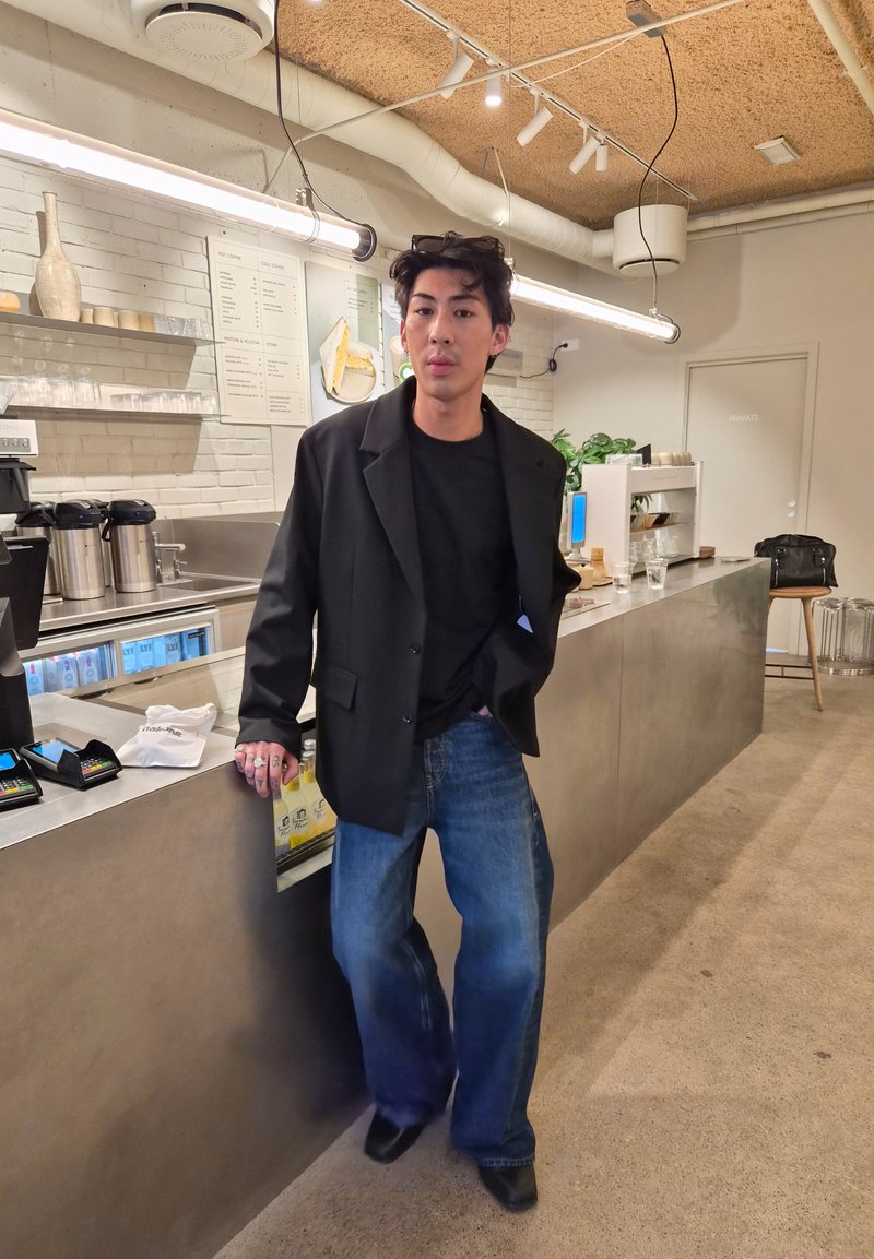 Personne avec un blazer noir et un jean appuyée sur le comptoir du café avec un menu, des machines à café et une décoration moderne à l’intérieur du café.