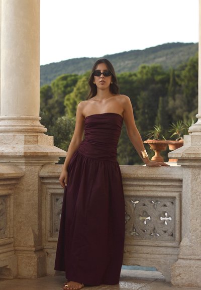 Vestido largo burdeos sin tirantes con corpiño fruncido y falda fluida. Combinado con gafas de sol negras y pulsera dorada, posando junto a una balaustrada de piedra.