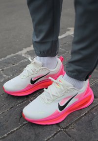 Chaussures de sport blanches avec des accents roses, dotées d'une tige en mesh texturé, d'un logo swoosh noir et d'une semelle rose rembourrée pour le soutien.