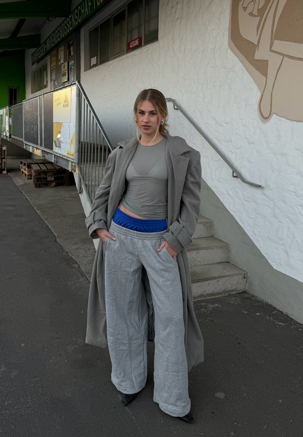 Model trägt einen hellgrauen Trenchcoat über einem figurbetonten grauen Oberteil und weiten grauen Sweatpants mit blauer elastischer Taille und steht im Freien.