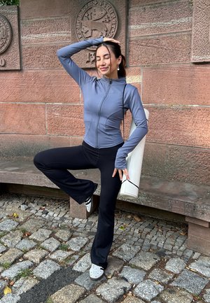 Chaqueta deportiva gris con cremallera y mangas largas, combinada con pantalones acampanados negros. Se sostiene un tapete de yoga blanco enrollado, con adoquines estampados debajo.