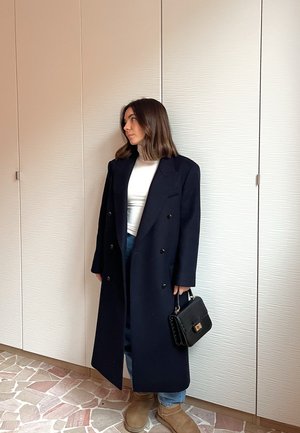 Mujer de pie en el interior vistiendo un abrigo largo negro, camiseta blanca, vaqueros azules, botas color canela y sosteniendo un bolso negro.