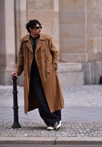 Dlhý béžový trenčkot s dvojradovým zapínaním, nosený cez tmavý sveter a voľné čierne nohavice s bielozelenými teniskami.