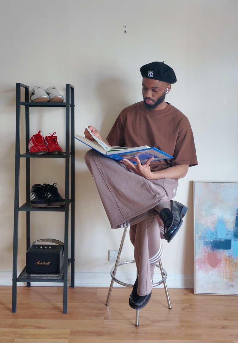 Homme portant un béret et des vêtements marron, assis sur un tabouret lisant un livre à côté d'un porte-chaussures et d'une peinture abstraite colorée sur le sol.
