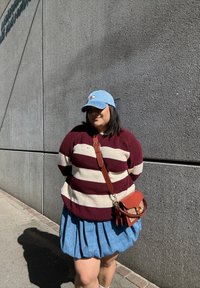 Maglione a righe marrone e crema, gonna di jeans, cappellino blu chiaro, borsa a tracolla marrone; persona in piedi contro un muro grigio testurizzato.