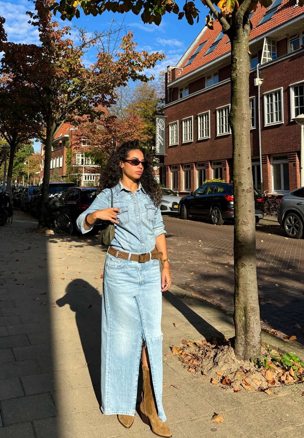 Ανοιχτό μπλε τζιν φόρεμα με άνοιγμα μπροστά, καφέ ζώνη και καφέ μπότες μέχρι το γόνατο. Το φόντο περιλαμβάνει τούβλινα κτίρια και φθινοπωρινά φύλλα.