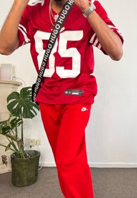 Camisola desportiva vermelha com números brancos, combinada com calças de treino vermelhas. Faixa preta com letras "HUGO", relógio prateado e uma planta verde ao fundo.