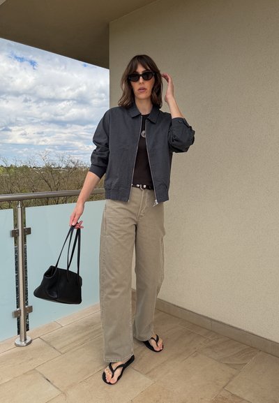 Mujer con chaqueta oscura, camiseta negra, pantalones beige de pierna ancha, gafas de sol y chanclas, sosteniendo un bolso negro en un balcón.