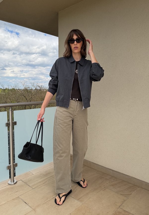Femme portant une veste sombre, un top noir, un pantalon beige à jambes larges, des lunettes de soleil et des tongs, tenant un sac à main noir sur un balcon.