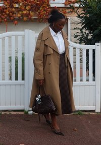 Długi, beżowy trencz z dwurzędowymi guzikami, noszony na białej koszuli i brązowej kratkowanej spódnicy. Nosi ciemną plecioną torebkę i szpilki.