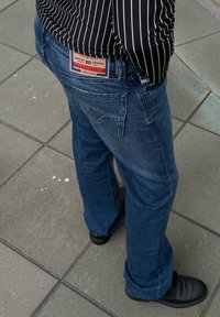 Dunkelblaue Jeans im geraden Schnitt mit Gesäßtaschen und sichtbarem Label; kombiniert mit schwarzen Schuhen auf einem gefliesten Untergrund.