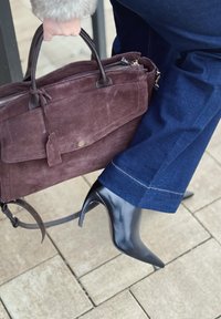Dunkelbraune Wildleder-Handtasche mit Reißverschluss und Klappentasche, gehalten von einer Hand, neben schwarzen spitzen High Heels und blauen Jeanshosen.