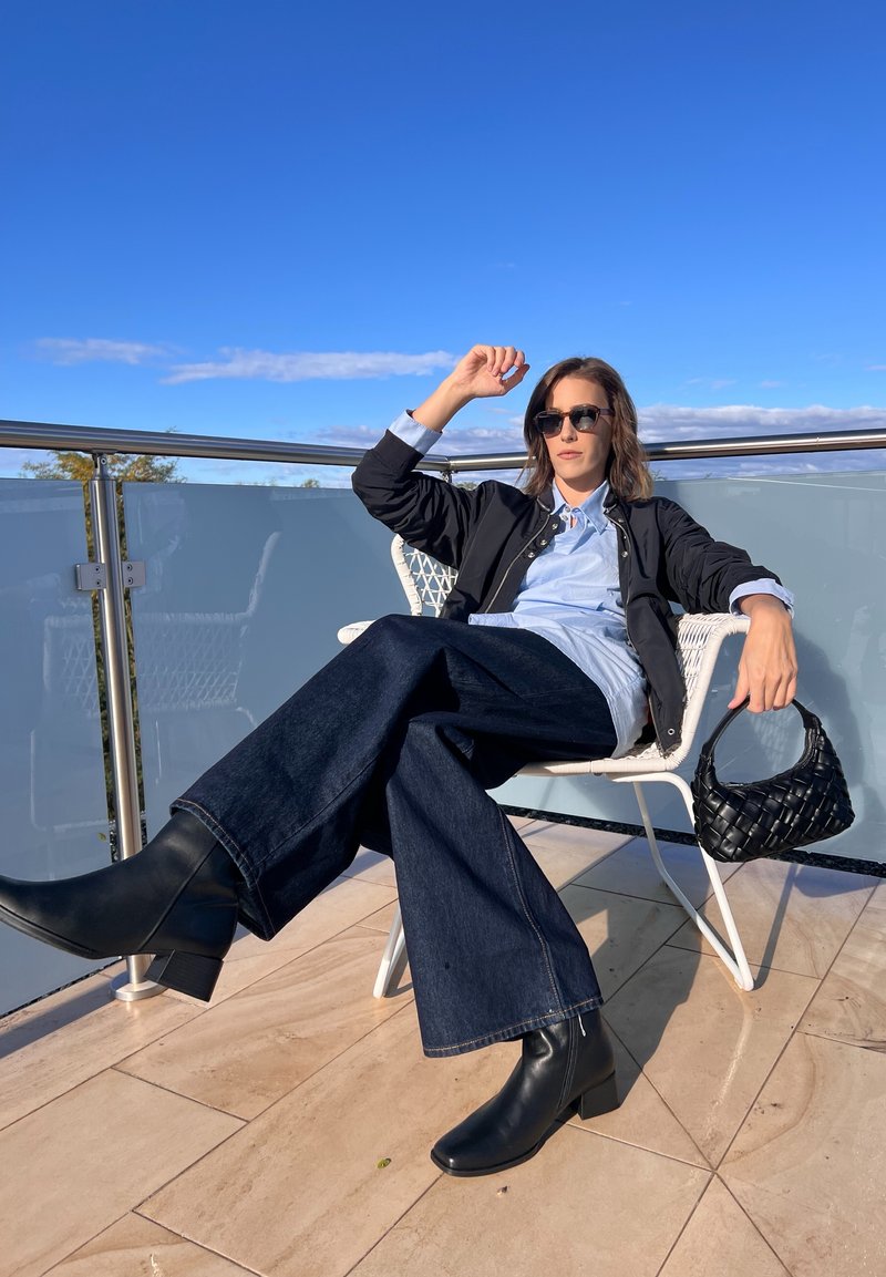 Μαυρή μπουφάν bomber πάνω από ένα ανοιχτό μπλε πουκάμισο, φαρδιά τζιν παντελόνια σε σκούρο ντένιμ, μαύρες ψηλές μπότες και μια πλεγμένη μαύρη τσάντα, καθισμένη σε μια καρέκλα.