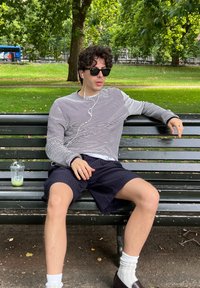 Stripete langermet skjorte, mørke shorts, solbriller og hvite sokker. Sitter på en benk med et klart plastglass i nærheten i en parkinnstilling.